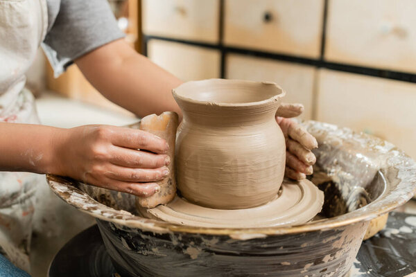 Cropped view of young female artisan in apron making clay vase with wooden scraper on pottery wheel in blurred ceramic workshop, clay shaping and forming process