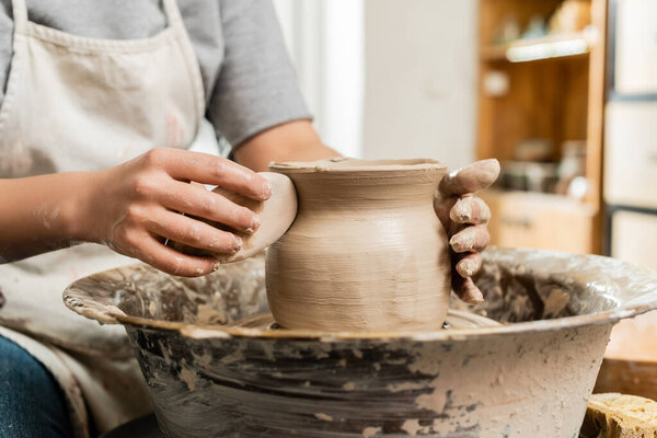 Cropped view of blurred young craftswoman in apron holding wooden scraper while making clay vase on pottery wheel in art studio at background, clay shaping and forming process