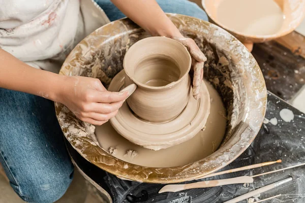 Top view of young female potter in apron making shape of clay vase with wooden tool on spinning pottery tool in ceramic workshop, clay shaping and forming process