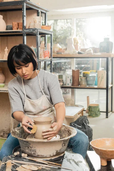 エプロンの若いアジアの女性職人は 湿ったスポンジで粘土花瓶の形を作り ぼやけたセラミックワークショップ 粘土形成と形成プロセスで回転陶器ホイールで作業します — ストック写真