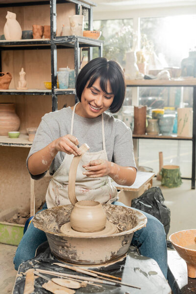 Cheerful young brunette asian artisan in apron holing clay while making vase on pottery wheel near tools and bowl in blurred ceramic workshop, artisanal pottery production and process