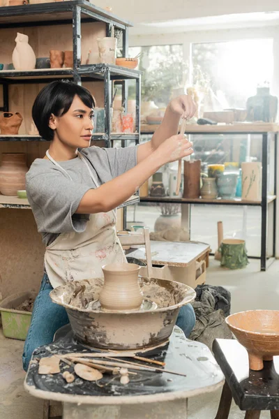 stock image Young brunette asian female potter in apron holding clay near vase on spinning pottery wheel near wooden tools and bowl in blurred ceramic workshop, artisanal pottery production and process