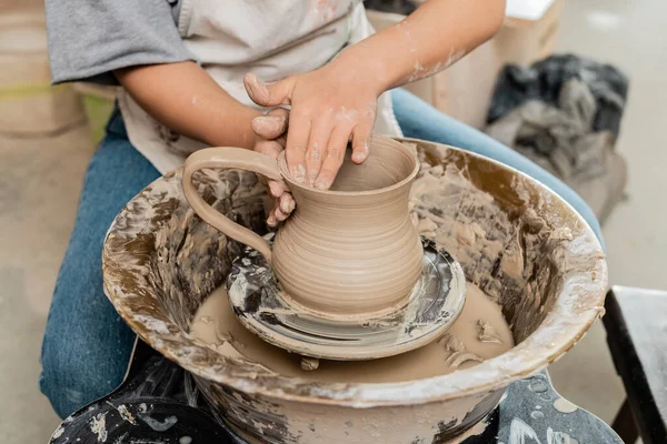 stock image Cropped view of young female artist in apron creating clay jug on pottery wheel on table while working in ceramic workshop, artisanal pottery production and process