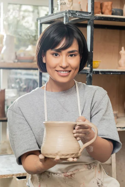 エプロンで若いブルネットのアジアの女性職人の肖像画と粘土のジャグを保持し 背景でぼやけたセラミックワークショップでカメラを見て 粘土の形成技術とプロセス — ストック写真