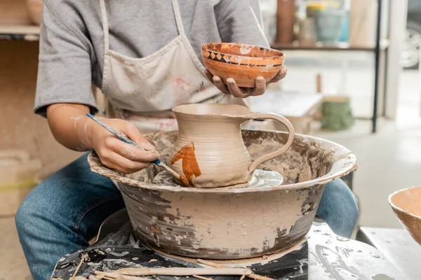 木製の道具の近くの陶器の車輪の上の粘土のジャグにエプロン塗装の若い女性アーティストの作物ビューぼやけたセラミックワークショップでテーブル 粘土成形技術とプロセス — ストック写真