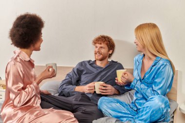 open relationship concept, polygamy, happy interracial women in pajamas holding cups of coffee and chatting with man, lovers, bisexual, understanding, three adults, cultural diversity, acceptance  clipart