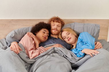 Açık ilişki, çok eşlilik, anlayış, battaniyenin altında uyuyan üç yetişkin, pijamalı kızıl saçlı adam ve çok kültürlü kadınlar, sevgililer, sabah, kültürel çeşitlilik, kabullenme, biseksüel kadınlar. 