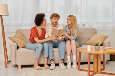diversity, open relationships, redhead man reading book to multicultural women in living room, non traditional lovers, non monogamy, romance and love, modern family and polygamy concept  clipart