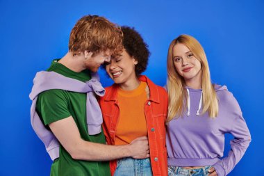 polyamory concept, cheerful redhead man hugging african american woman near blonde lover on blue background, studio photography, cultural diversity, polygamy, modern family, understanding  clipart