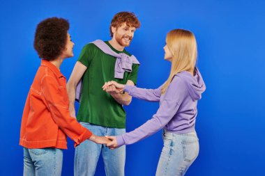 polyamorous relationship concept, polygamy lovers, young man and multicultural women holding hands on blue background, studio shot, denim fashion, love triangle, bonding and acceptance  clipart