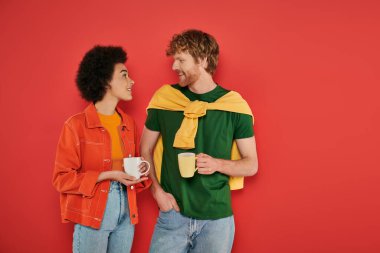 multicultural couple holding cups of coffee on coral background, morning routine, cultural diversity, vibrant colors, stylish outfits, interracial lovers holding mugs and having conversation  clipart