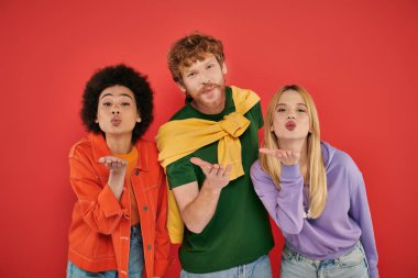 Açık ilişki, çok eşlilik, mercan arka planında hava öpücüğü veren üç ırklar arası sevgili, kültürel çeşitlilik, çok eşli, mutlu, çok ırklı insanlar kameraya bakıyor. 