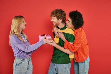 open relationship, blonde woman presenting gift box to redhead man near african american bisexual lover, polyamorous people on coral background, holiday, festive occasions, present  clipart