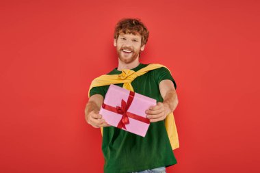 holiday, cheerful redhead man with beard posing in casual attire on coral background, holding gift box, festive occasions, wrapped present, fashion and trend, urban style, happiness  clipart