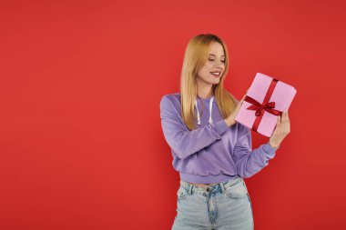 holiday, cheerfulness, blonde woman in casual attire holding present on coral background, smiling, vibrant colors, wrapped gift box, attractive and stylish, festive occasions  clipart