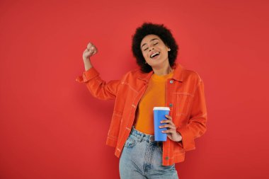 takeaway drink, cheerful african american woman in casual attire holding paper cup on coral background, vibrant colors, attractive and stylish, coffee to go, excited and inspired  clipart