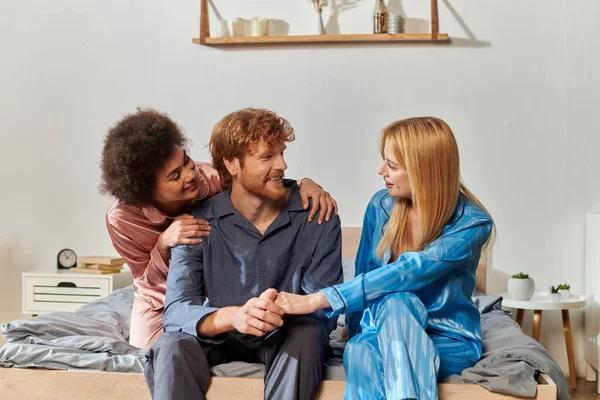 open relationship concept, three adults, redhead man holding hands with blonde woman, multicultural people in pajamas sitting on bed at home, cultural diversity, acceptance, bisexual