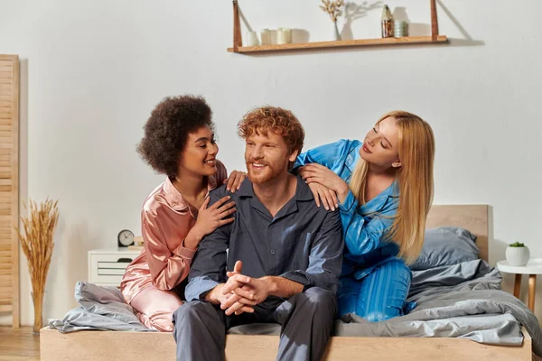 stock image polyamory concept, open relationship, polygamy, three adults, happy redhead man and multicultural women in pajamas sitting on bed at home, cultural diversity, acceptance, bisexual 