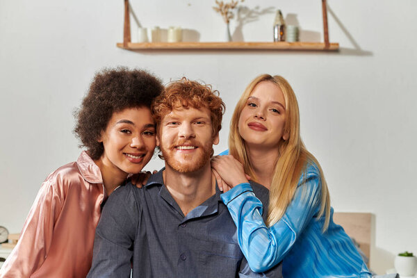 polyamory concept, open relationship, polygamy, portrait of three adults, happy redhead man and multicultural women in pajamas looking at camera, cultural diversity, acceptance, bisexual 