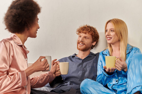 open relationship concept, polygamy, happy man chatting with interracial women in pajamas, holding cups of coffee, lovers, bisexual, understanding, three adults, cultural diversity, acceptance 