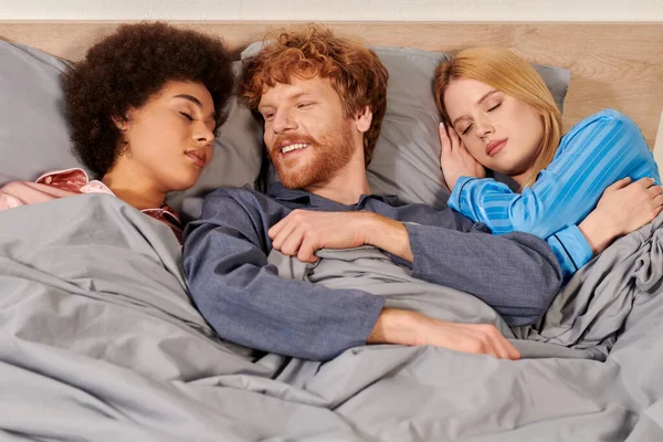 non traditional relationship, polygamy, three adults, happy redhead man waking up near multicultural women, threesome, cultural diversity, acceptance, bisexual