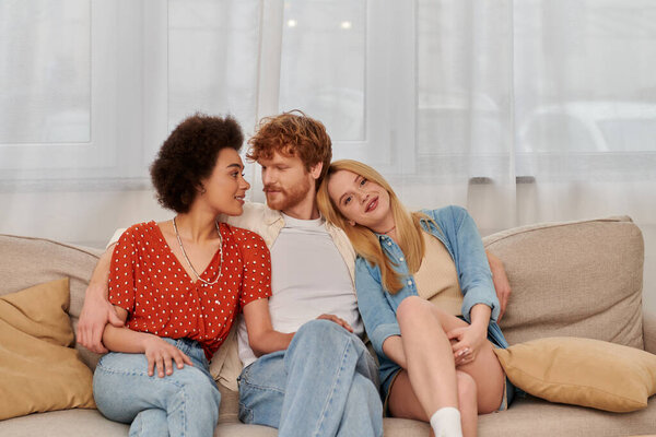 modern family, polygamy concept, freedom in relationship, cultural diversity, redhead man sitting with multicultural women on couch in living room, polyamorous lifestyle, non traditional 