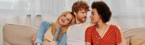 modern family, polygamy concept, freedom in relationship, cultural diversity, redhead man sitting with multicultural women on couch in living room, polyamorous lifestyle, non traditional, banner 