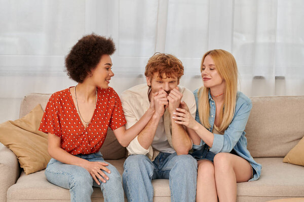 polyamorous relationship, cultural diversity, redhead man sitting on couch with happy multiracial female lovers, freedom and acceptance, love triangle, three people in open relationship 