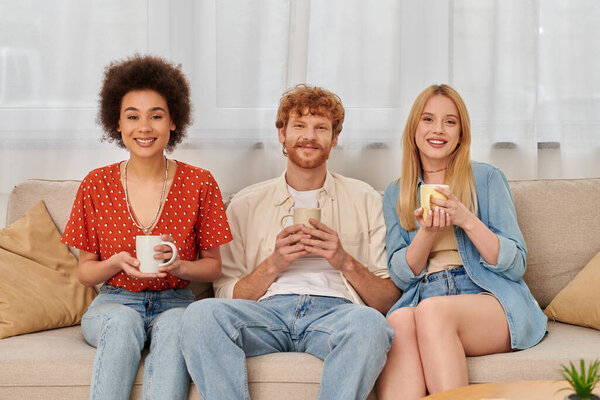 polyamorous concept, relationship diversity, happy polygamy lovers sitting on couch and holding cups of coffee, looking at camera, interracial man and women in living room, bisexual and polygamy 