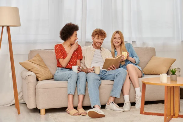 stock image open relationships, polygamy, intelligent redhead man reading book to multicultural women in living room, modern family, hobby and leisure, polyamorous family, cups of coffee 