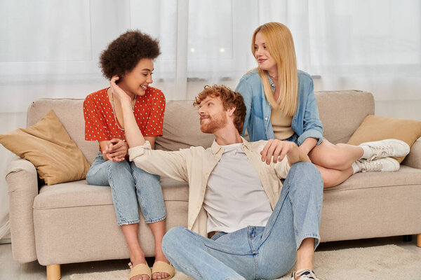 love triangle concept, diversity in relationships and culture, non monogamy, happy redhead man sitting near multicultural women in living room, lovers, modern family, acceptance, open relationship 