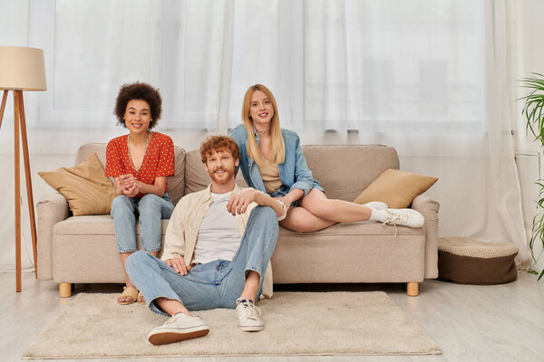 love triangle concept, diversity in relationships and culture, non monogamy, happy redhead man and multicultural women looking at camera in living room, lovers, acceptance, open relationship 