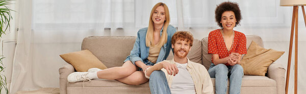 love triangle concept, diversity in relationships and culture, non monogamy, happy redhead man and multicultural women looking at camera in living room, lovers, acceptance, open relationship, banner 