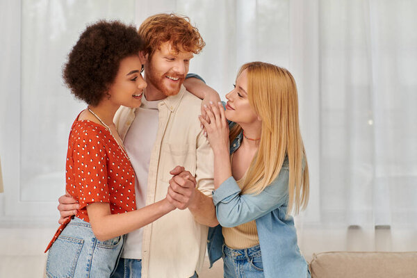 alternative family, happy multicultural women near redhead man in living room, interracial lovers, cultural diversity, freedom in relationships, love triangle, bisexual and free spirited people 