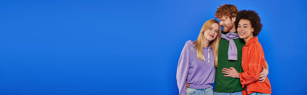 polyamory concept, cheerful multicultural women looking at camera and hugging with redhead man on blue background, studio shot, cultural diversity, polygamy, young lovers in casual attire, banner 
