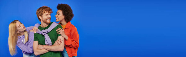 alternative relationships, polyamory, three people, young redhead man and beautiful multiracial women on blue background vibrant colors, casual clothes, stylish attire, modern family, banner 
