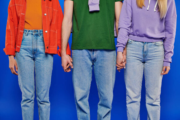 alternative family, cropped view of three polygamy people, young man and women holding hands on blue background, studio shot, vibrant clothes, polyamory, modern relationships 