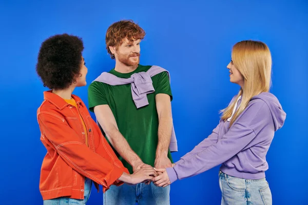多色関係の概念 一夫多妻制の人々 青の背景に手を取り合って若い男と多文化の女性 スタジオショット デニムファッション 愛の三角形 結合と愛 — ストック写真