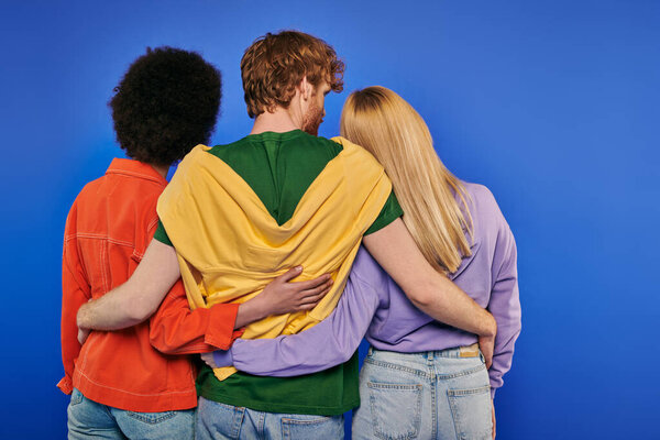 love triangle, polyamory lovers, back view of young redhead man hugging multiracial women on blue background, studio shot, vibrant colors, casual clothes, stylish attire, modern family 