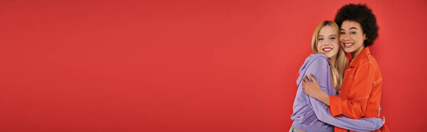 cultural diversity, cheerful multicultural women hugging while looking at camera on coral background, blonde and brunette, diverse friends, sisterhood, friendship goals, studio shot, banner 