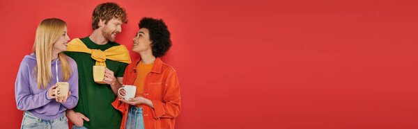 polygamy concept, young multicultural women and redhead man holding cups on coral background, studio shot, vibrant colors, morning routine, coffee, happy polyamorous lovers, banner 