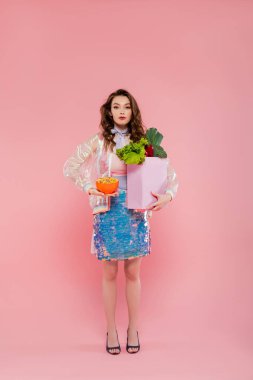 Ev hanımı konsepti, sebzeli market çantası taşıyan çekici bir kadın pembe arka planda dalgalı saçlı bir model kavramsal fotoğrafçılık, ev işleri, şık bir eş. 