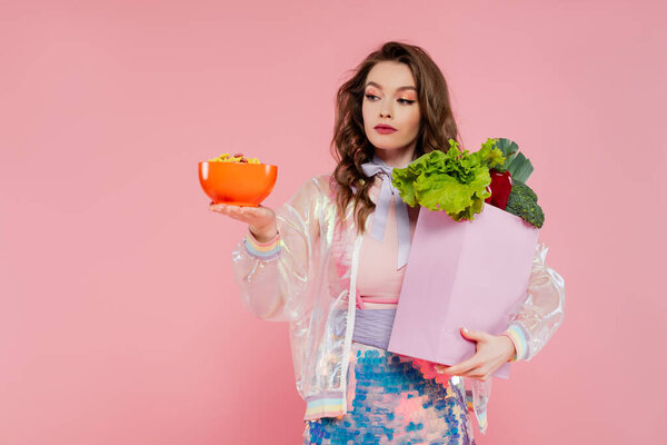 housewife concept, attractive young woman carrying grocery bag with vegetables and bowl with corn flakes, model with wavy hair on pink background, conceptual photography, home duties, breakfast 