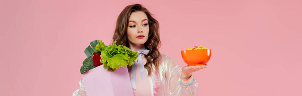 housewife concept, attractive young woman carrying grocery bag with vegetables and bowl with corn flakes, model with wavy hair on pink background, conceptual photography, home duties, banner