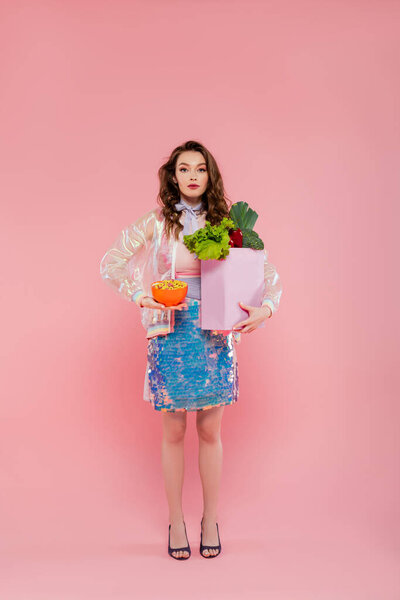 housewife concept, doll like, attractive young woman carrying grocery bag with vegetables, model with wavy hair on pink background, conceptual photography, home duties, stylish wife 