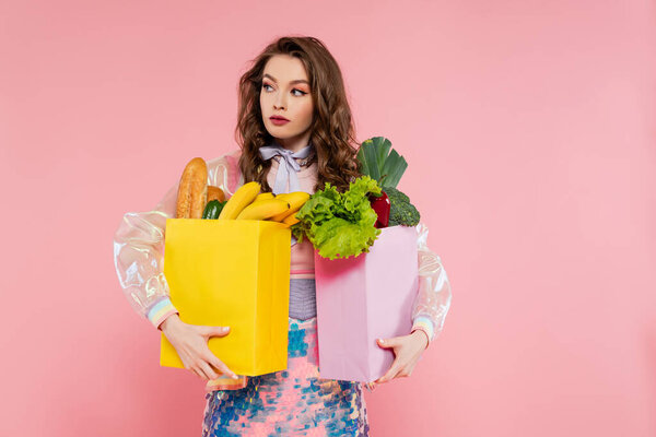 housewife concept, attractive young woman carrying grocery bags with vegetables and bananas, model with wavy hair on pink background, conceptual photography, home duties, stylish wife 