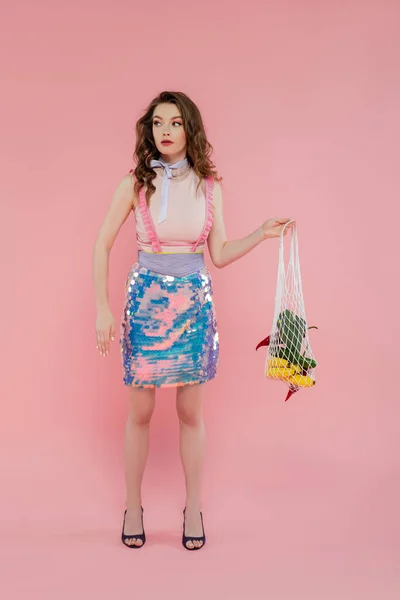 stock image housewife concept, beautiful young woman posing and gesturing like a doll, holding reusable mesh bag with groceries, trendy wife doing daily house duties, standing on pink background, eco-friendly 