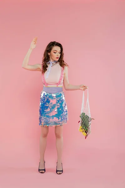 stock image housewife concept, attractive young woman posing and gesturing like a doll, holding reusable mesh bag with groceries, stylish wife doing daily house duties, standing on pink background 