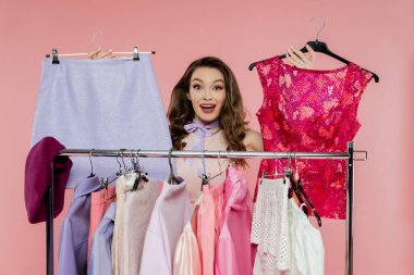 consumerism, amazed young woman with brunette wavy hair standing near rack with clothes, showing trendy outfit, wardrobe selection concept, beautiful model looking at camera on pink background  clipart