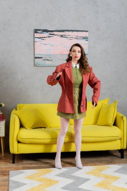 konsept fotoğrafçılık, oyuncak bebek gibi davranan kadın, doğal olmayan hareketler yapan ve sarı kanepenin yanında duran, iyi giyimli ve güzel, modern ev içi, rol yapma oyunu, bebek hayatı 